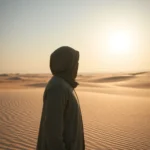 Person in einem Wüstenumhang steht im Sand bei Sonnenuntergang.