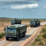 Drei militärische LKW mit Tarnmuster auf einer staubigen Straße in einer hügeligen Landschaft.